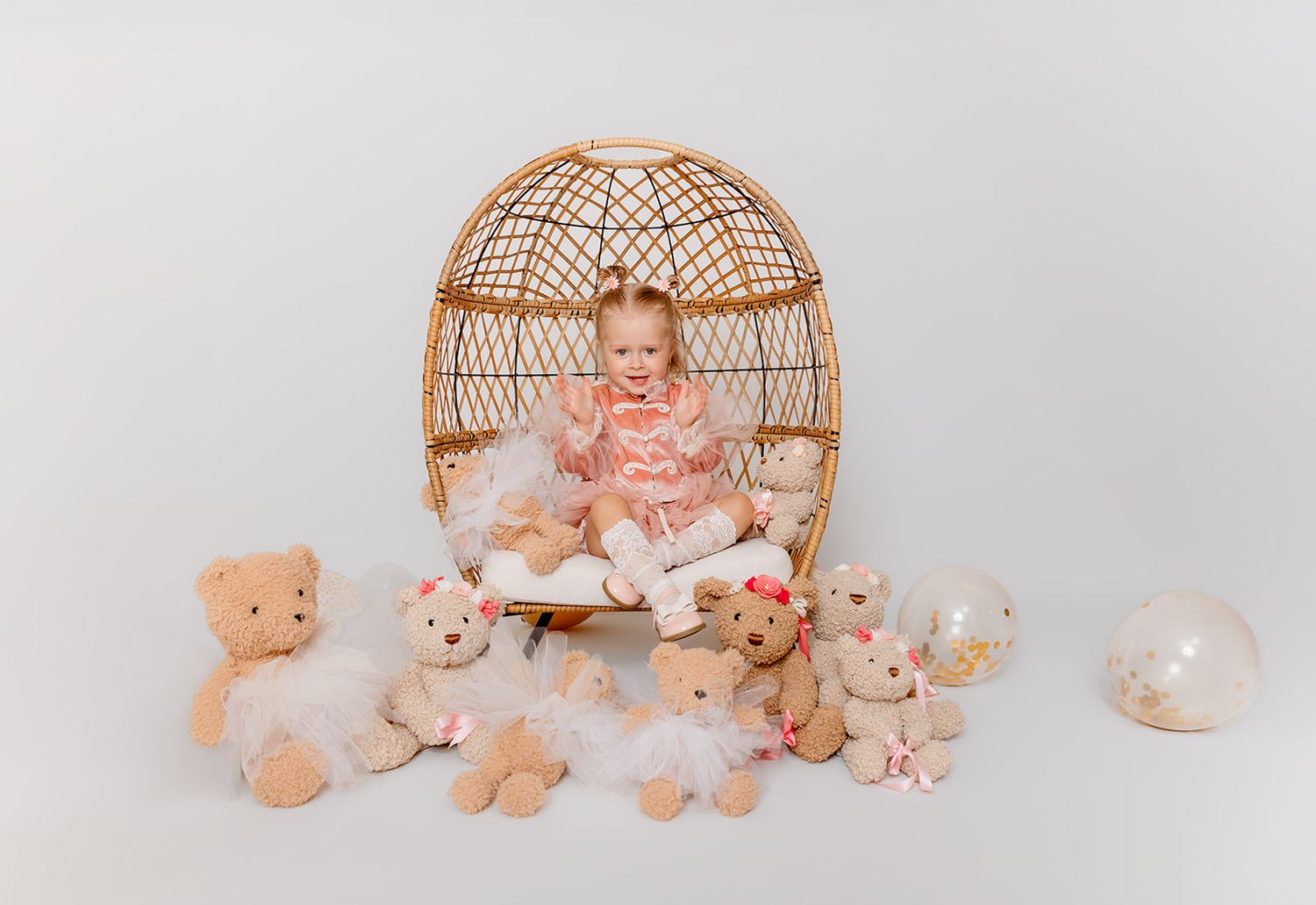 Baby girl in pink dress sits in wicker egg chair surrounded by teddy bears with tutus on white background.