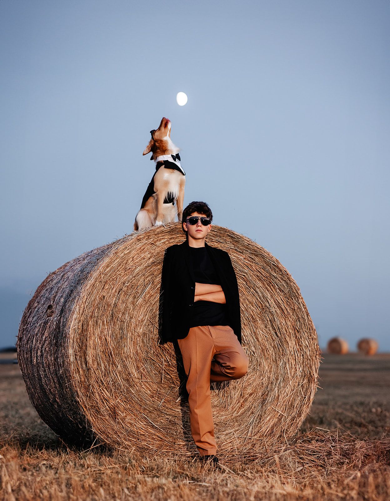 Person in black sweater and orange pants leaning on hay bale with beagle dog on top under blue sky.