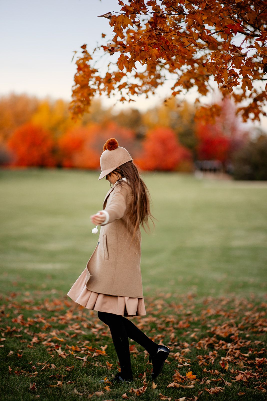 A young girl in a beige coat and hat walks among fallen autumn leaves in a colorful fall park.