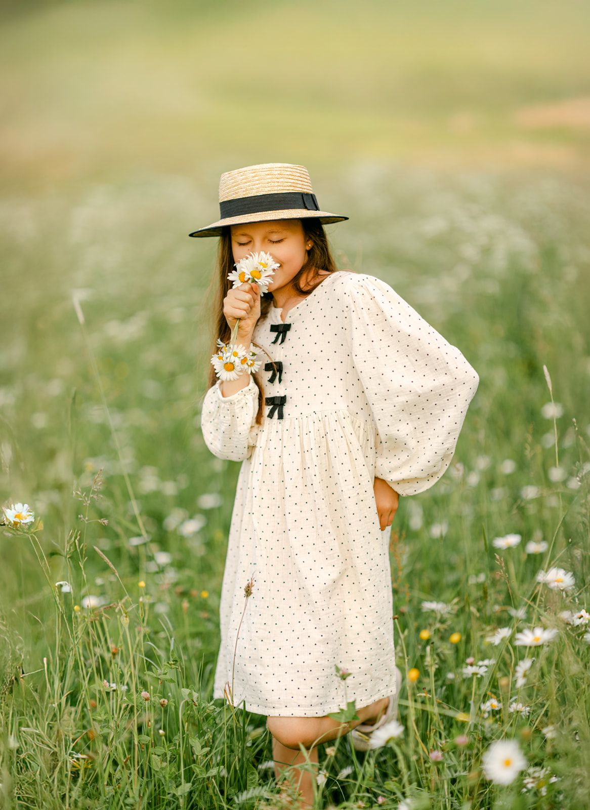 Young girl in white polka dot dress and straw hat smells daisies in a sunny meadow.