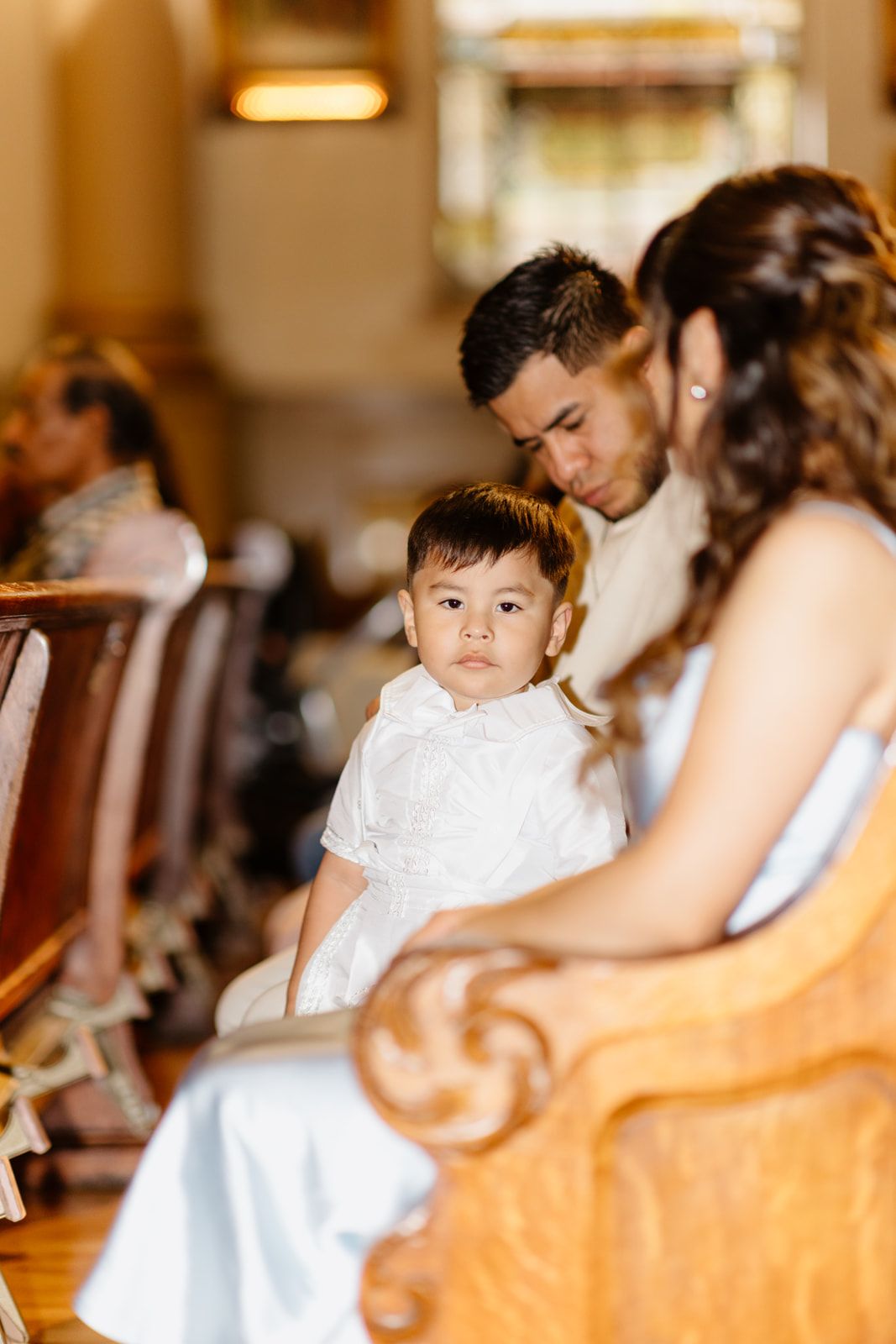 A young child in white sits on a parent's lap during a church baptism ceremony.