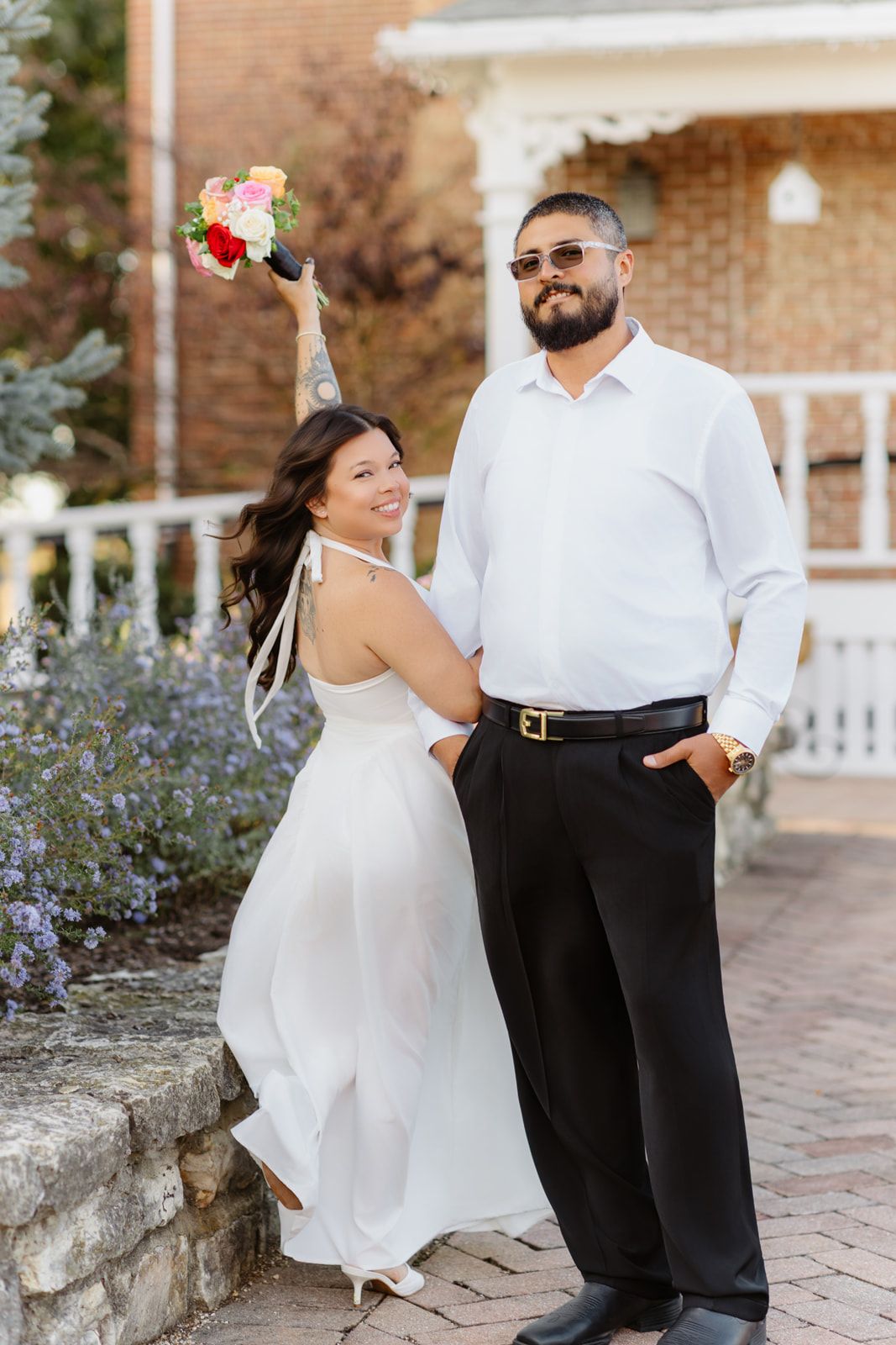 Bride in white dress raises bouquet while groom in white shirt and sunglasses stands beside her on brick path.