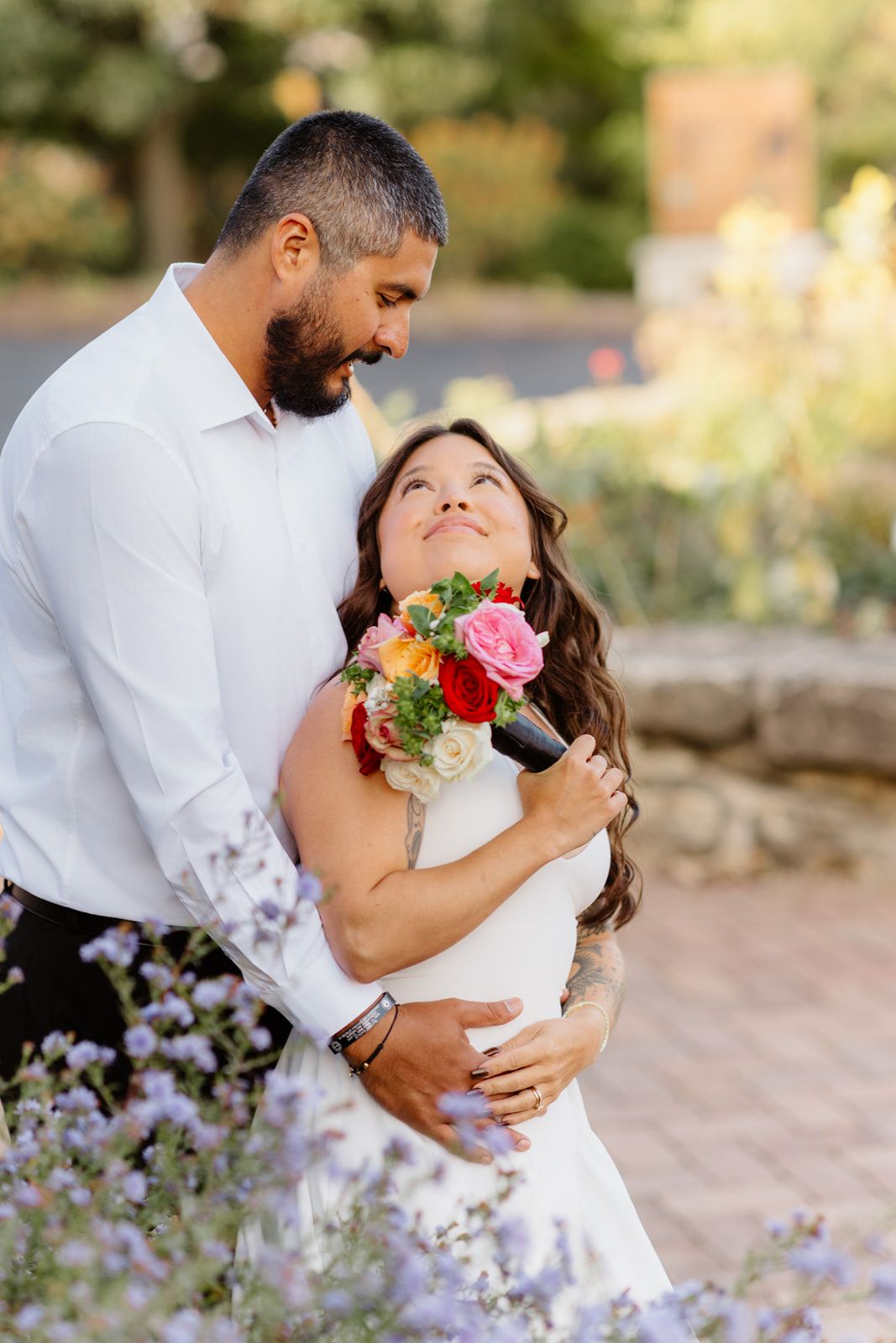 Bride and groom embrace outdoors, she holds colorful bouquet gazing up at him in wedding attire.
