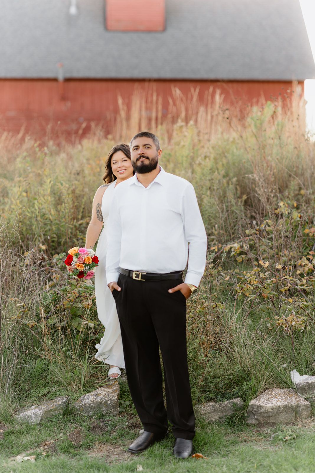 Bride and groom pose outdoors near a red barn, she holds a colorful bouquet while standing behind him.