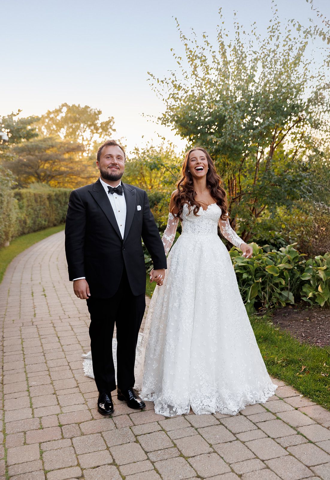 Laughing bride in lace gown holds hands with groom on a garden pathway during golden hour.