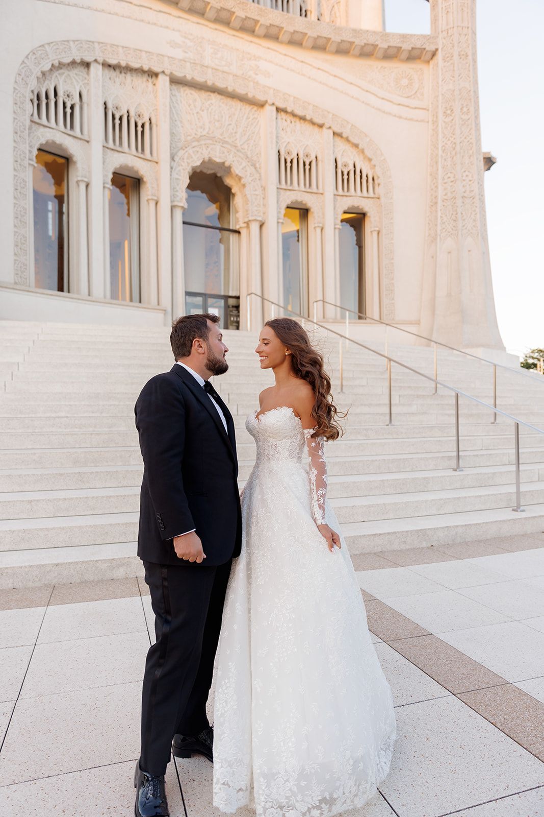 Bride and groom share a romantic moment on steps of ornate white building, she in lace gown, he in black tuxedo.