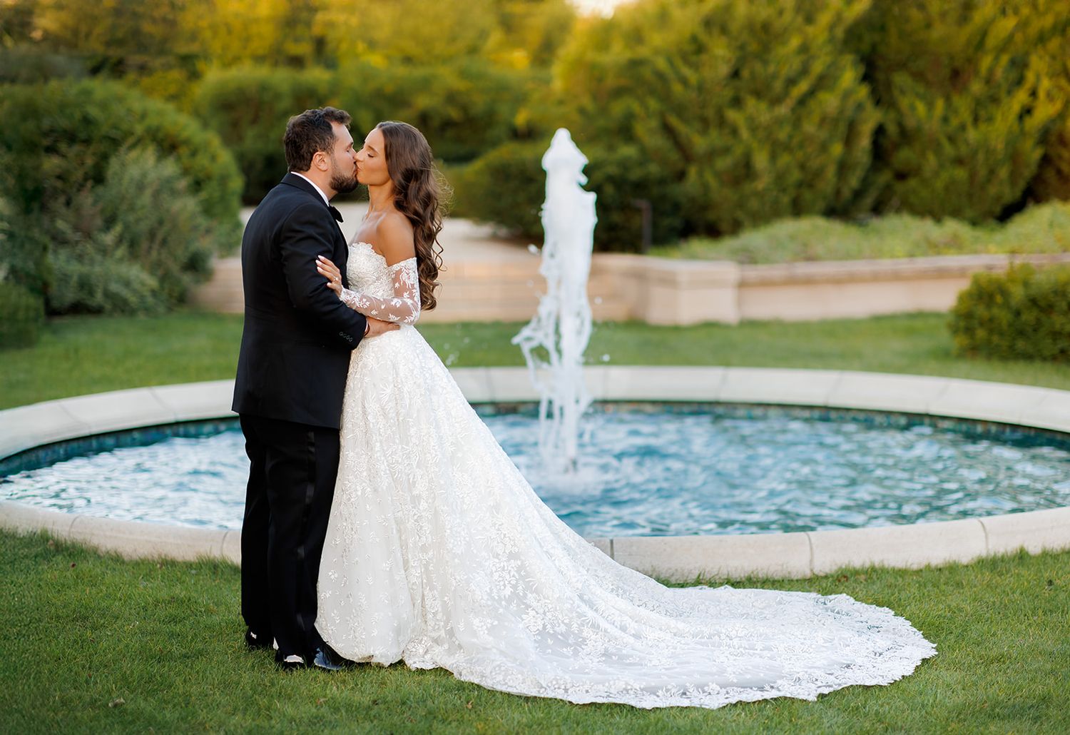 Bride and groom share a romantic kiss in elegant wedding attire beside a garden fountain on their wedding day.