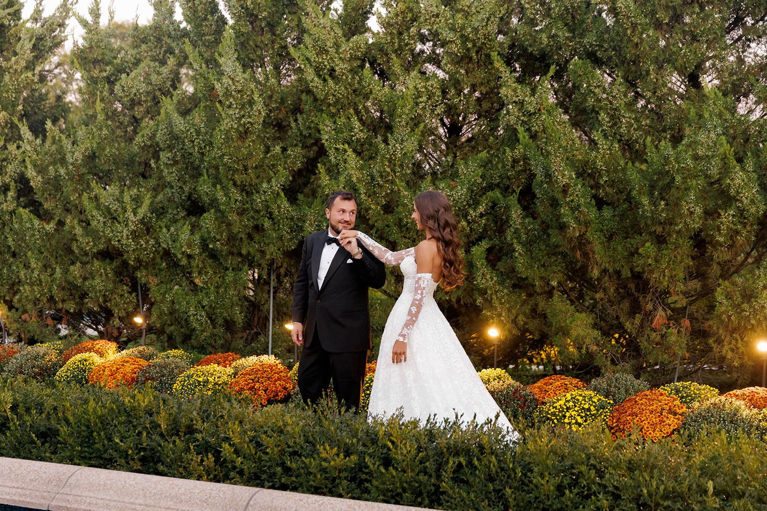 Bride and groom share a romantic moment in a lush garden with yellow chrysanthemums and evergreen trees.