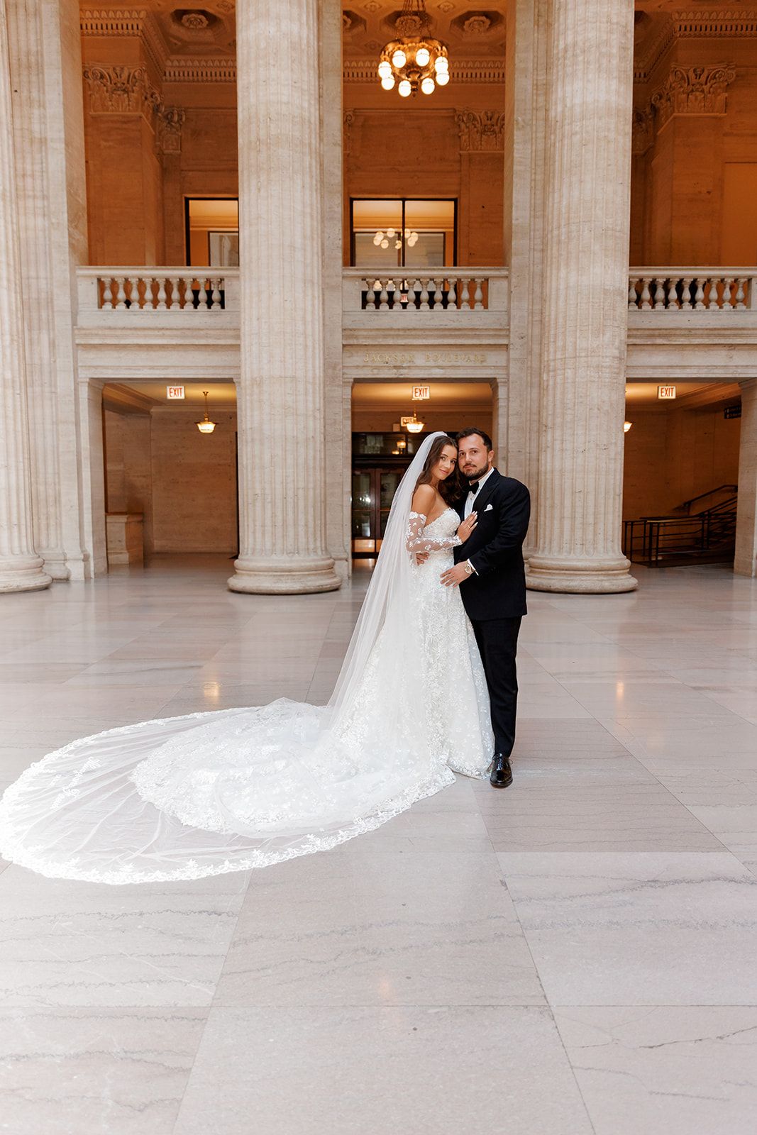 Bride and groom pose elegantly in a grand marble hall with tall columns, her lace cathedral veil trailing beautifully.