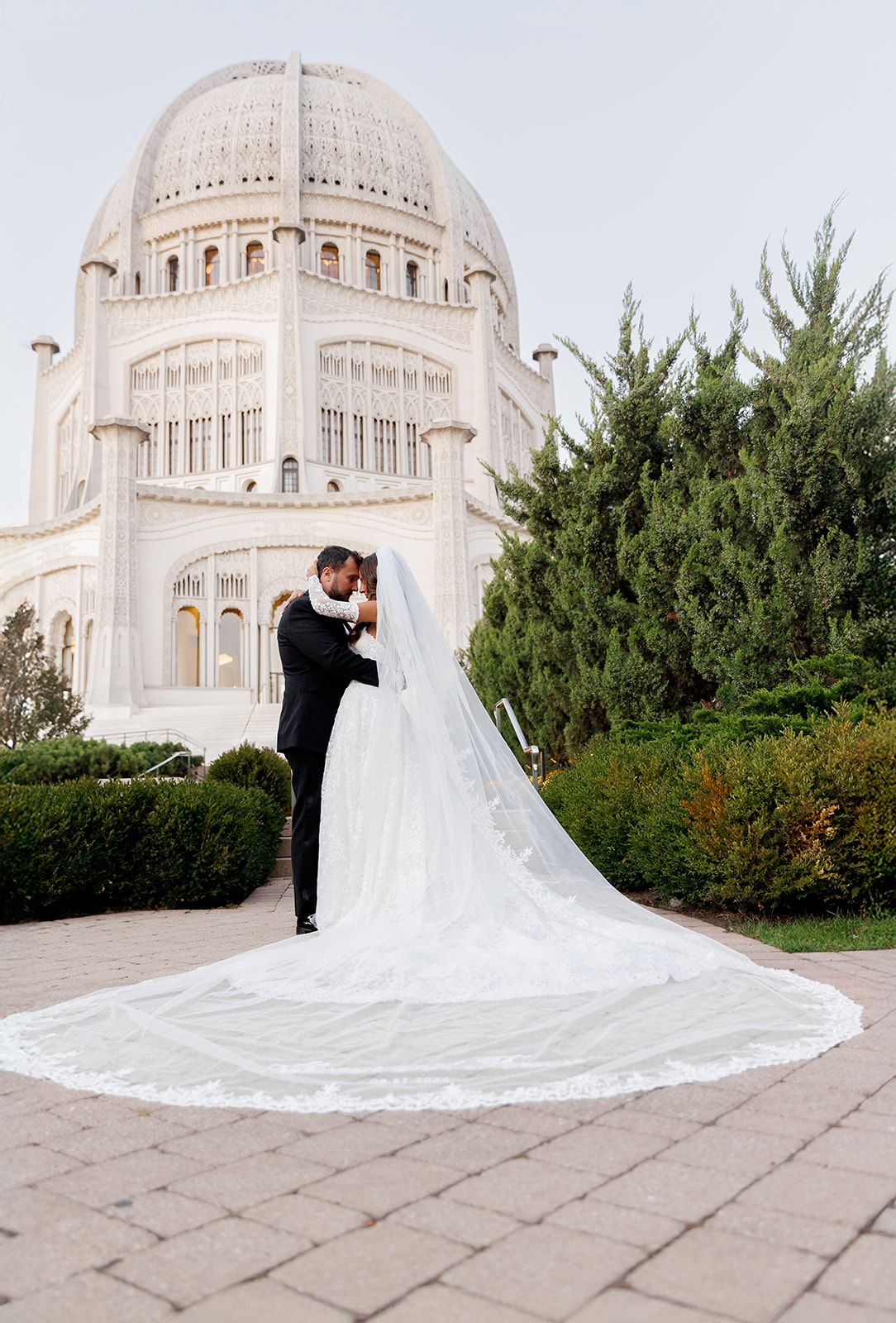 Bride and groom embrace before the Bahá'í Temple in Wilmette, IL, her cathedral veil spreading elegantly on the stone plaza.