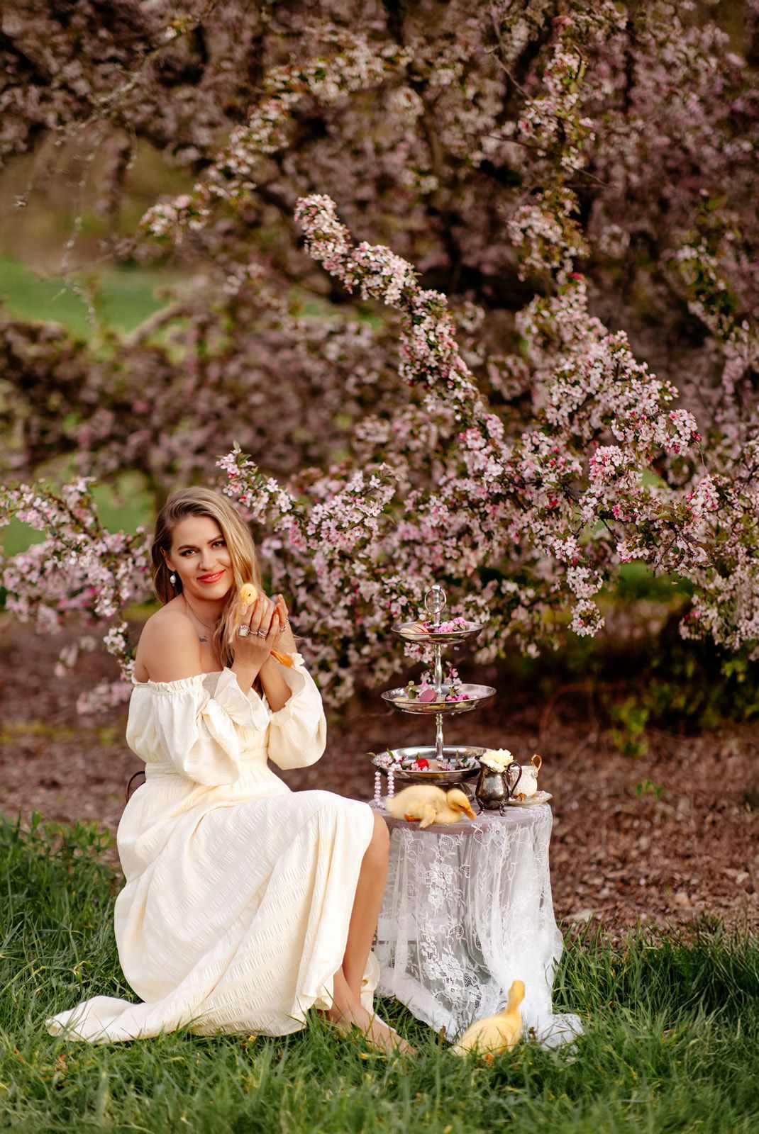 Woman in white off-shoulder dress sits by blooming cherry tree with tiered cake stand at spring picnic.