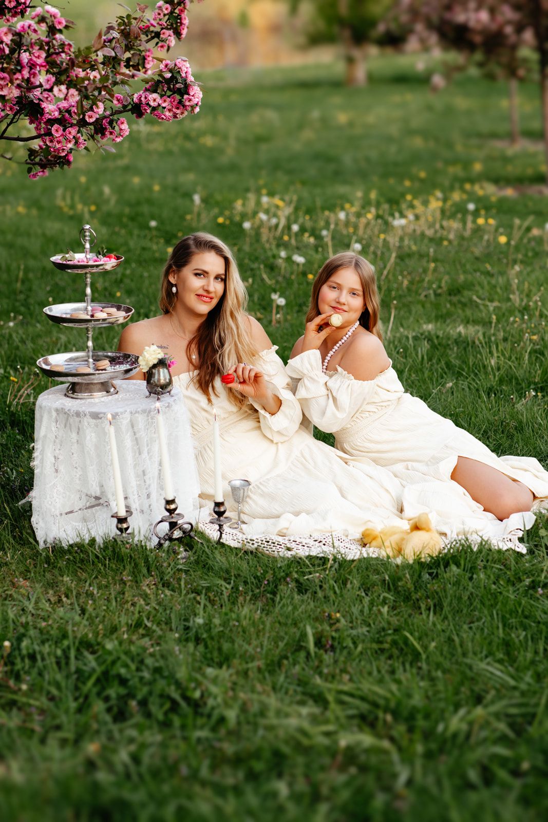 A woman and girl in white dresses enjoy a garden picnic with ducklings, candles, and a tiered tray under blooming trees.
