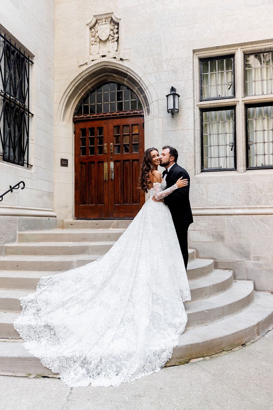 Bride in elegant lace gown with long train stands with groom in black tux on stone steps of historic building.