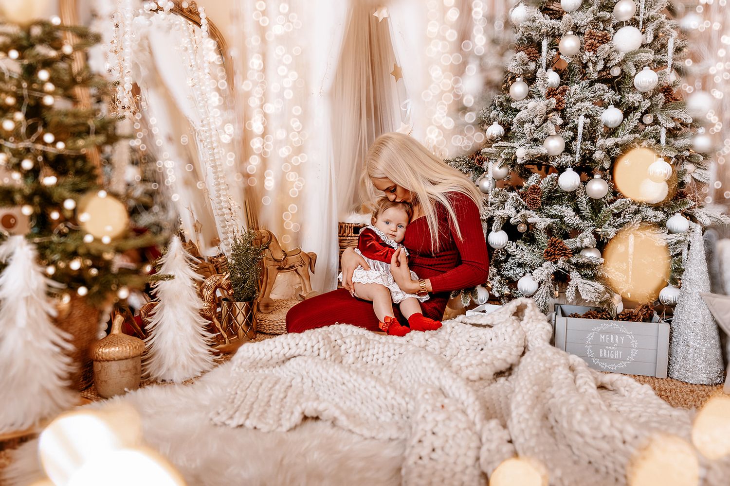Mother in red dress holds baby near decorated Christmas trees with white knit blanket and glowing holiday lights.