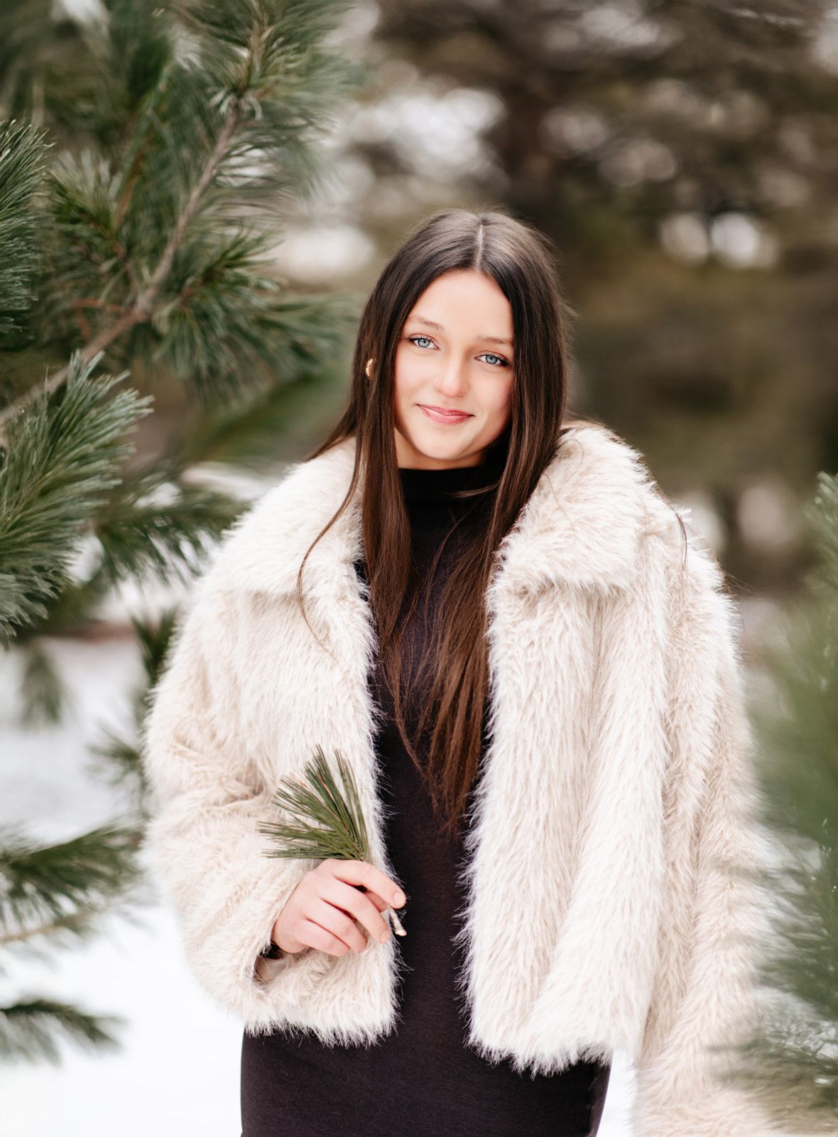 Smiling brunette teen in cream fur coat holding pine sprig beside snowy evergreen tree.