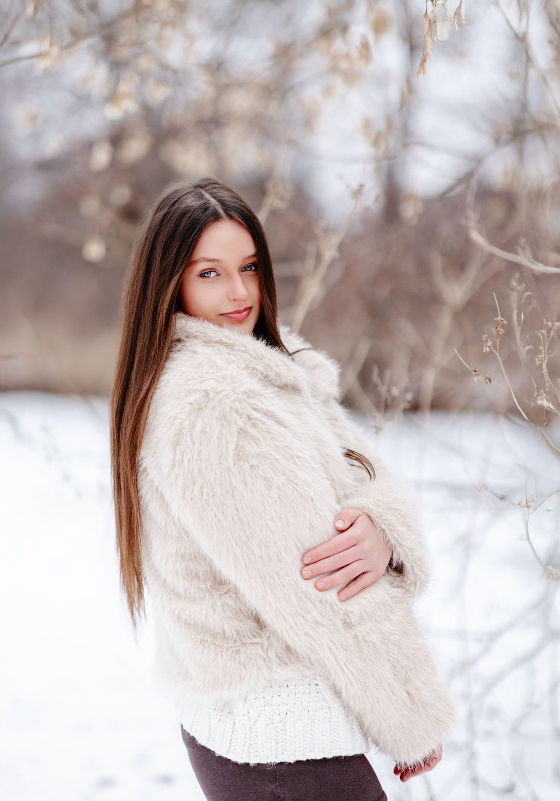 Young woman with long brown hair in a fluffy white coat poses outdoors in a snowy winter landscape.