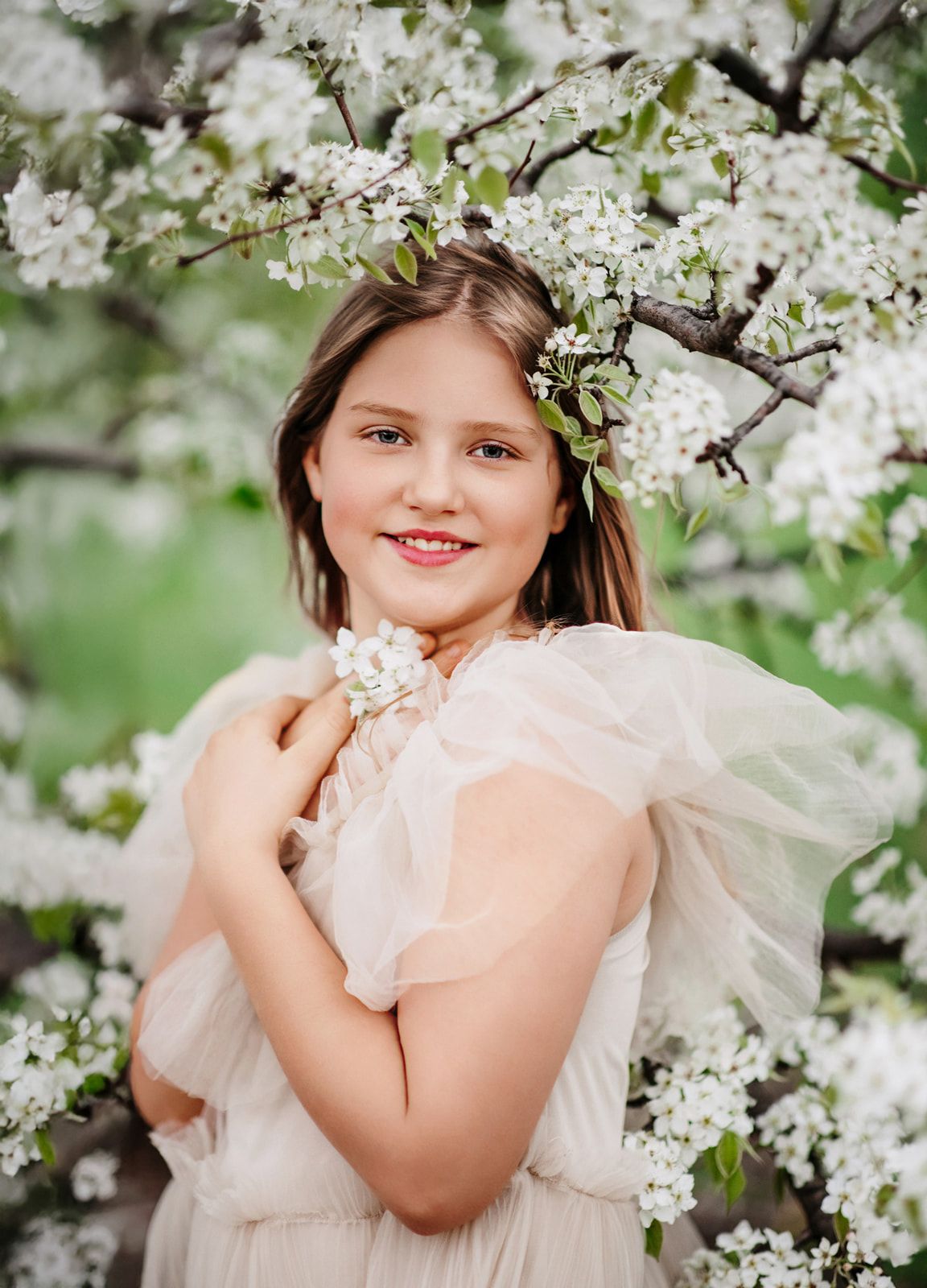 Young girl in cream tulle dress smiling among white spring blossoms, holding flowers in a dreamy outdoor portrait.