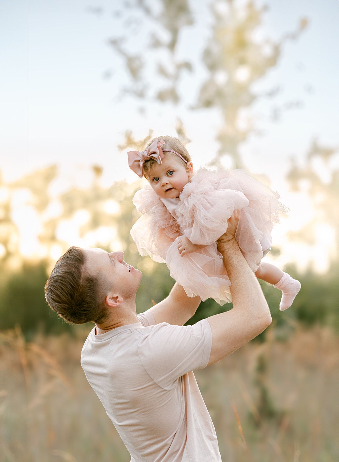 A father lifts his smiling baby girl in a pink tutu dress outdoors in a golden sunlit field.