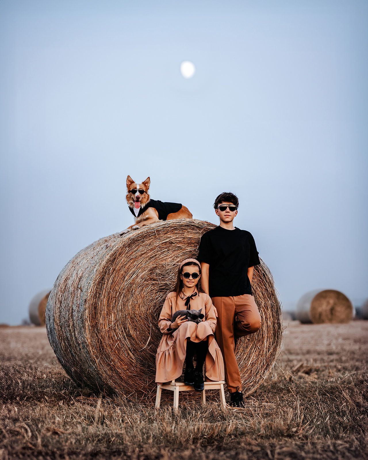 Two teens with sunglasses pose by a large hay bale in a field with a German Shepherd dog under full moon.