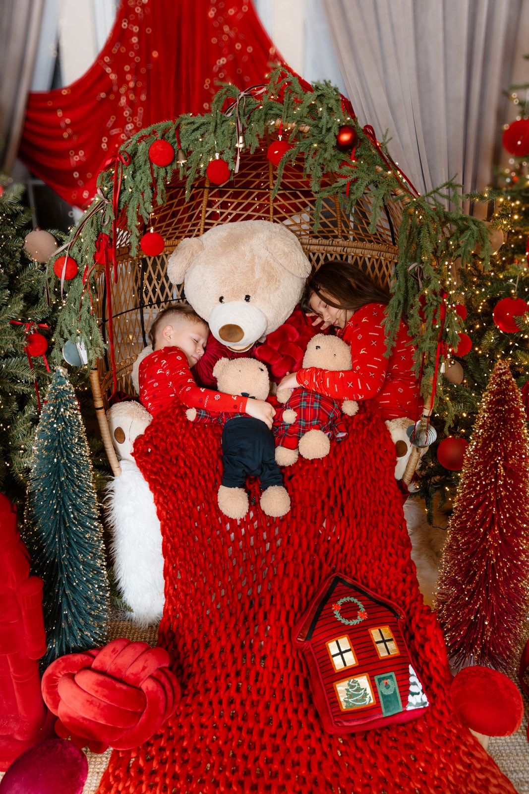 Two children sleeping in a wicker chair with a teddy bear amid festive Christmas decorations and red knit blanket.