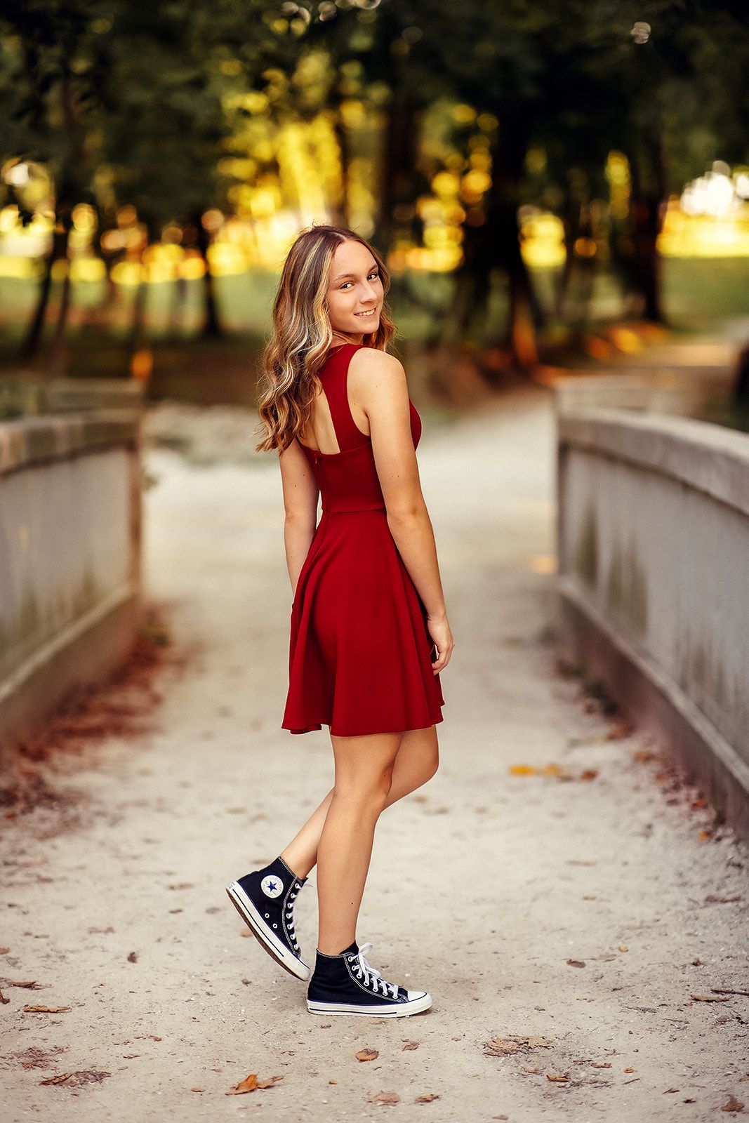 Young woman in red dress and black Converse sneakers poses on a sandy bridge path in a park.