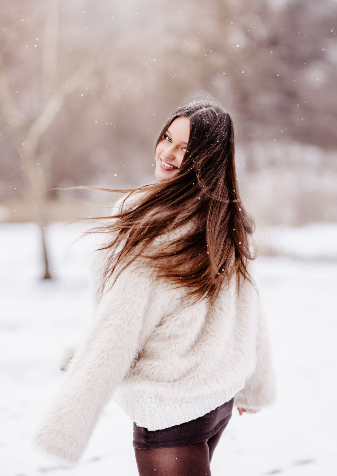 A smiling young woman with long brown hair twirls in a snowy winter park wearing a cozy white knit sweater.