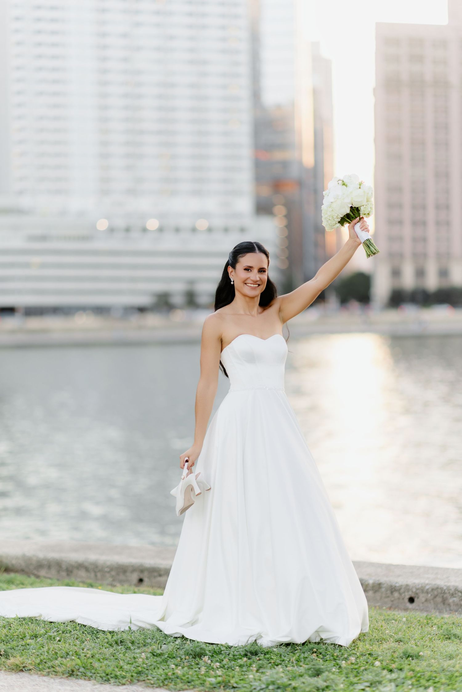 Smiling bride and bearded groom pose together by city river at golden hour, she holding white bouquet.