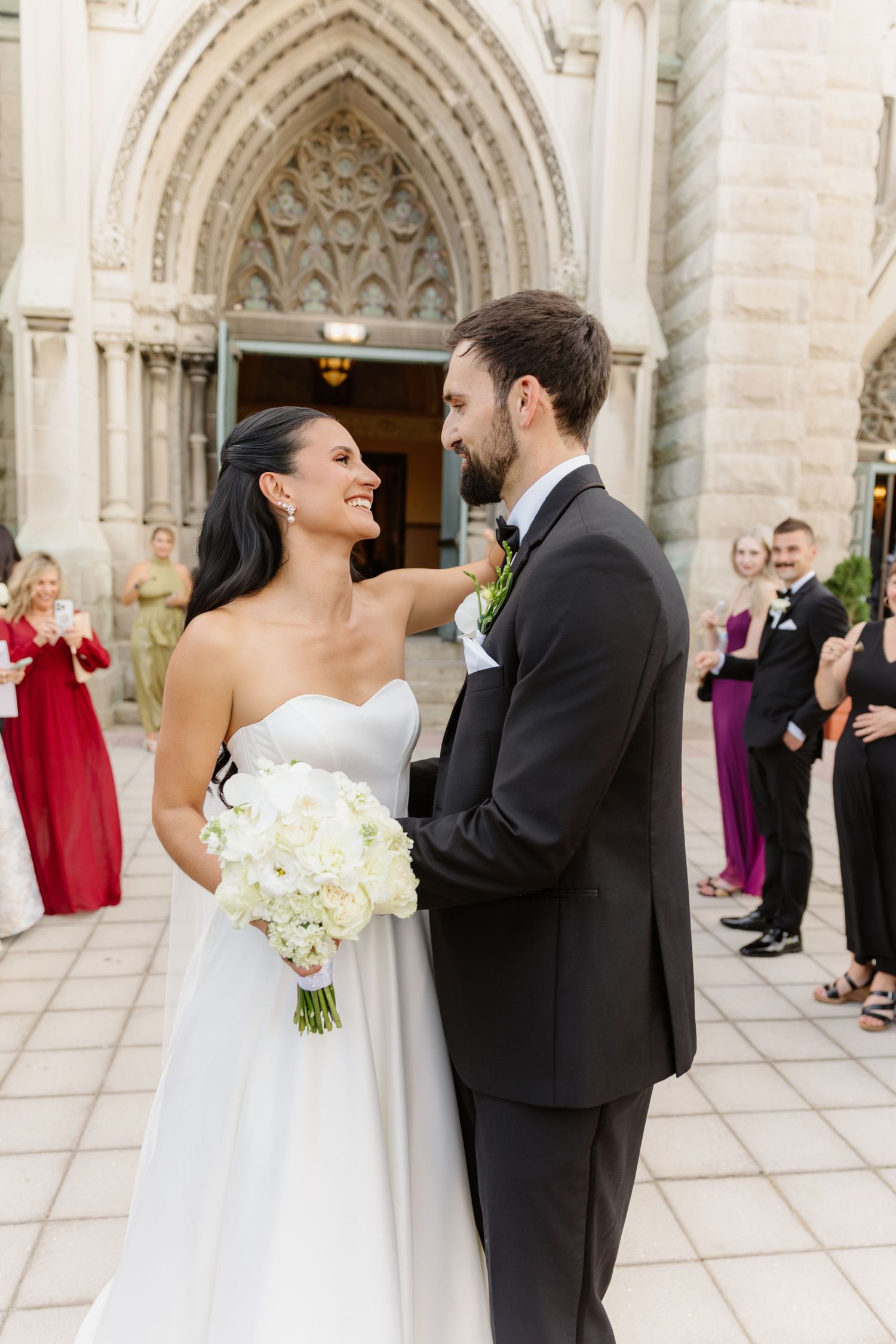Bride in strapless gown holds white bouquet smiling at bearded groom outside church.