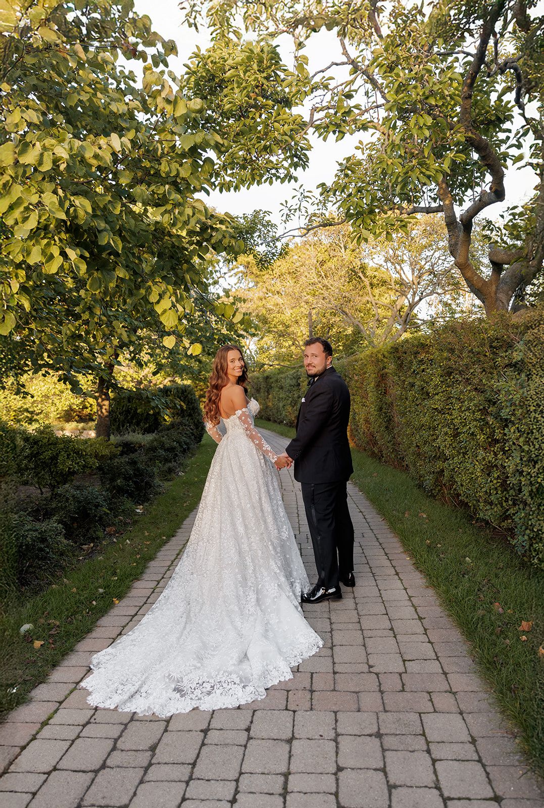 Wedding couple holding hands on a brick garden path, bride wearing long-sleeve lace gown with elegant train.