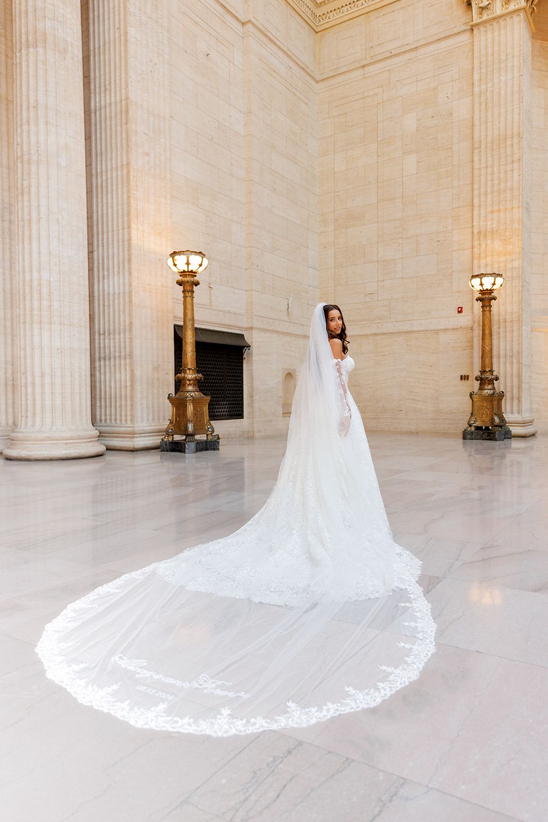 Bride in a white gown with an elegant lace cathedral veil spread across marble floors between gold columns.