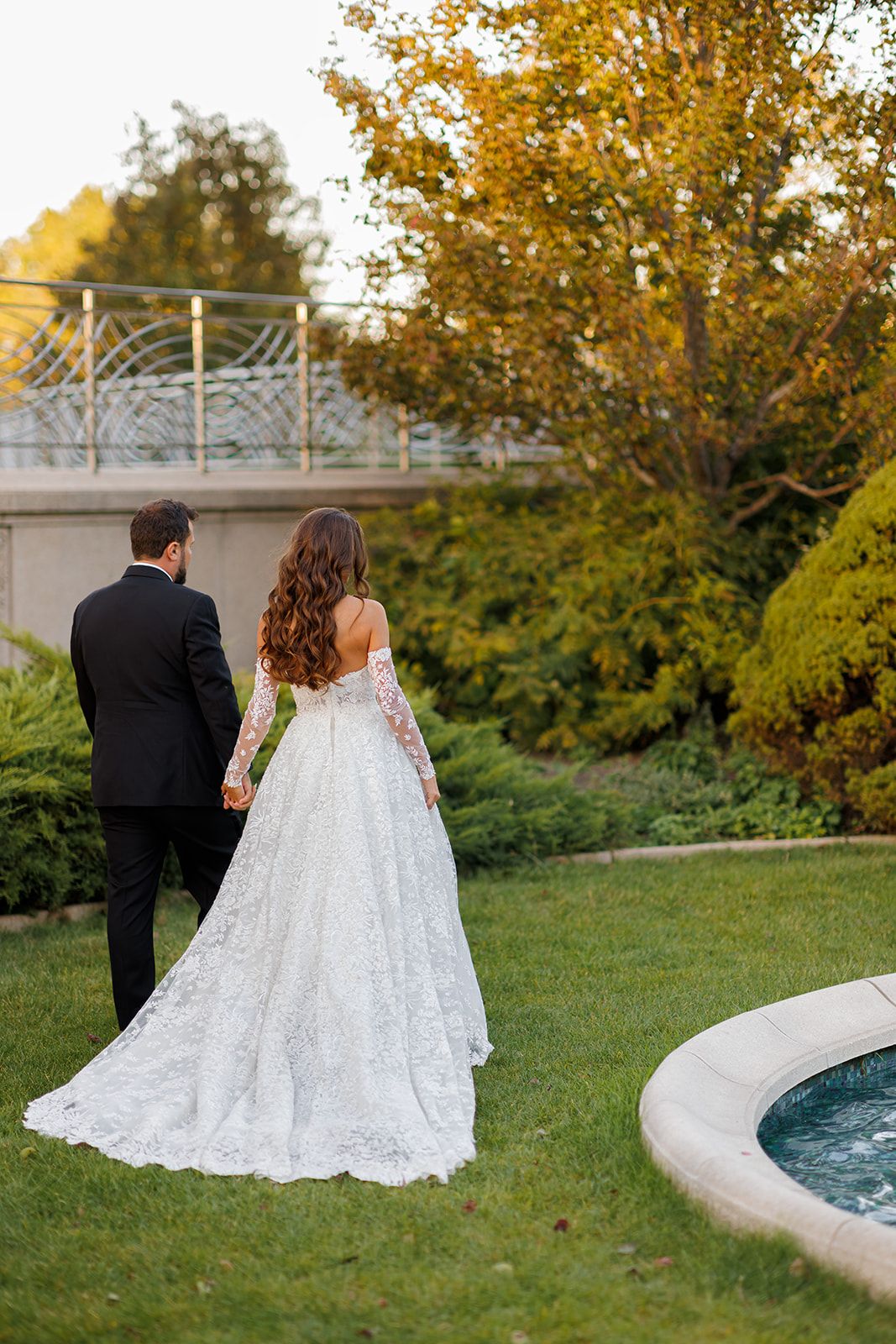 Bride in lace ball gown holds hands with groom in black tuxedo walking through lush garden near fountain.