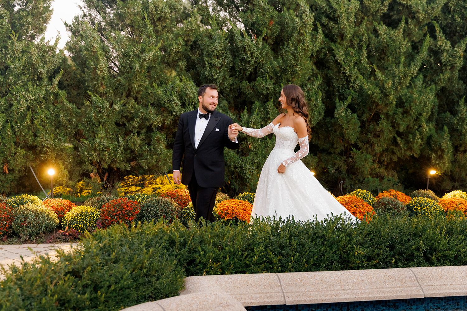 Bride and groom holding hands walking through lush garden with colorful fall flowers at elegant outdoor wedding venue.