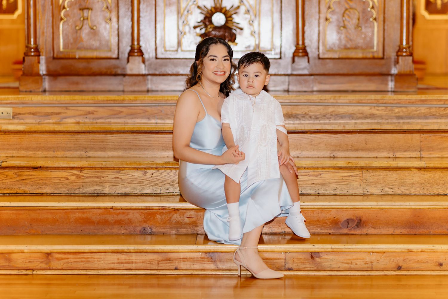 A woman in a light blue dress sits on ornate wooden stairs with a young child in white clothing.