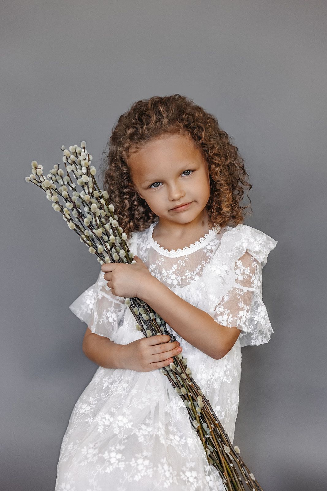 Little girl with curly hair in white lace dress holding pussy willow branches on gray background.