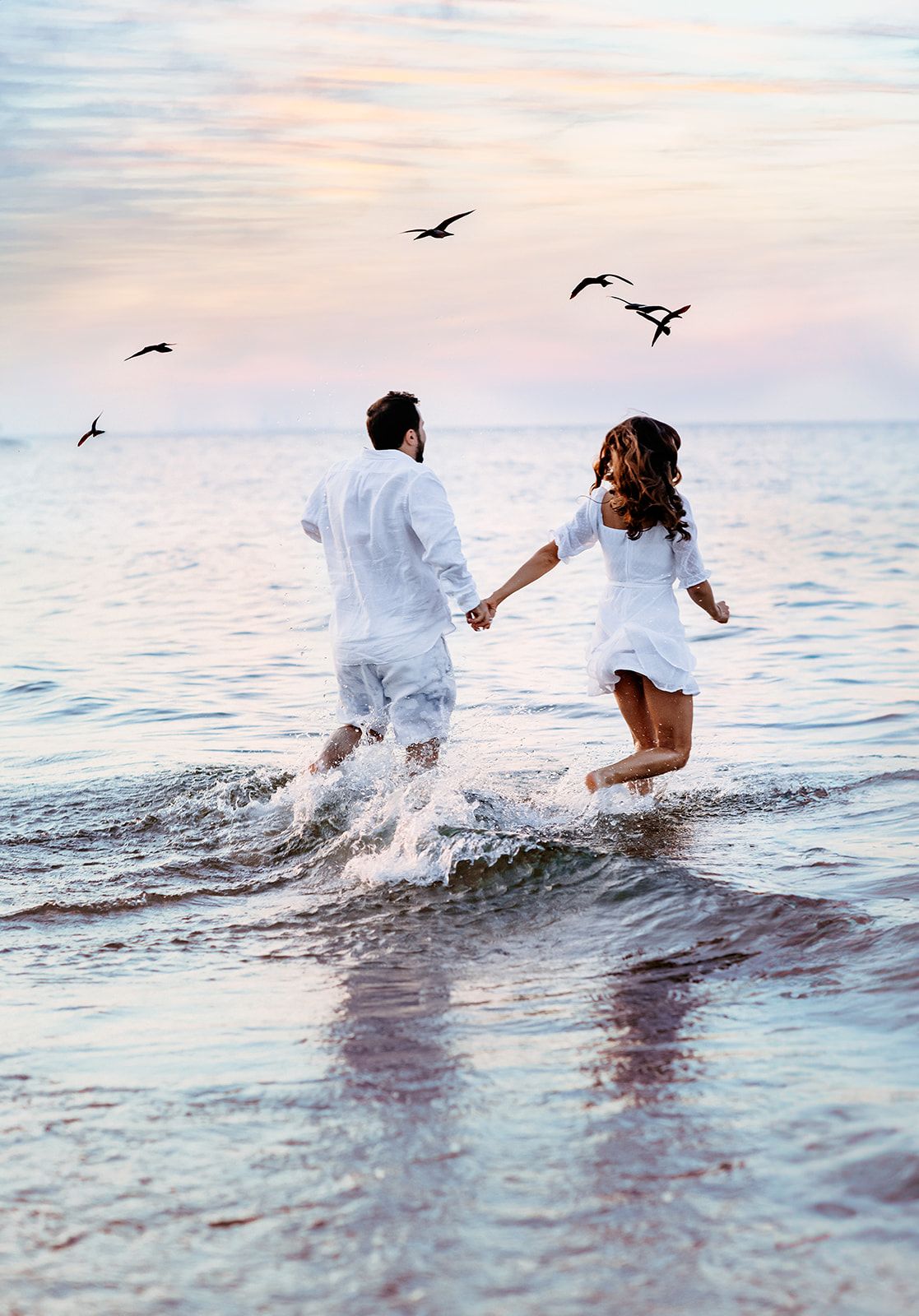 Carefree couple in white holding hands running through ocean waves at dusk with birds flying overhead.