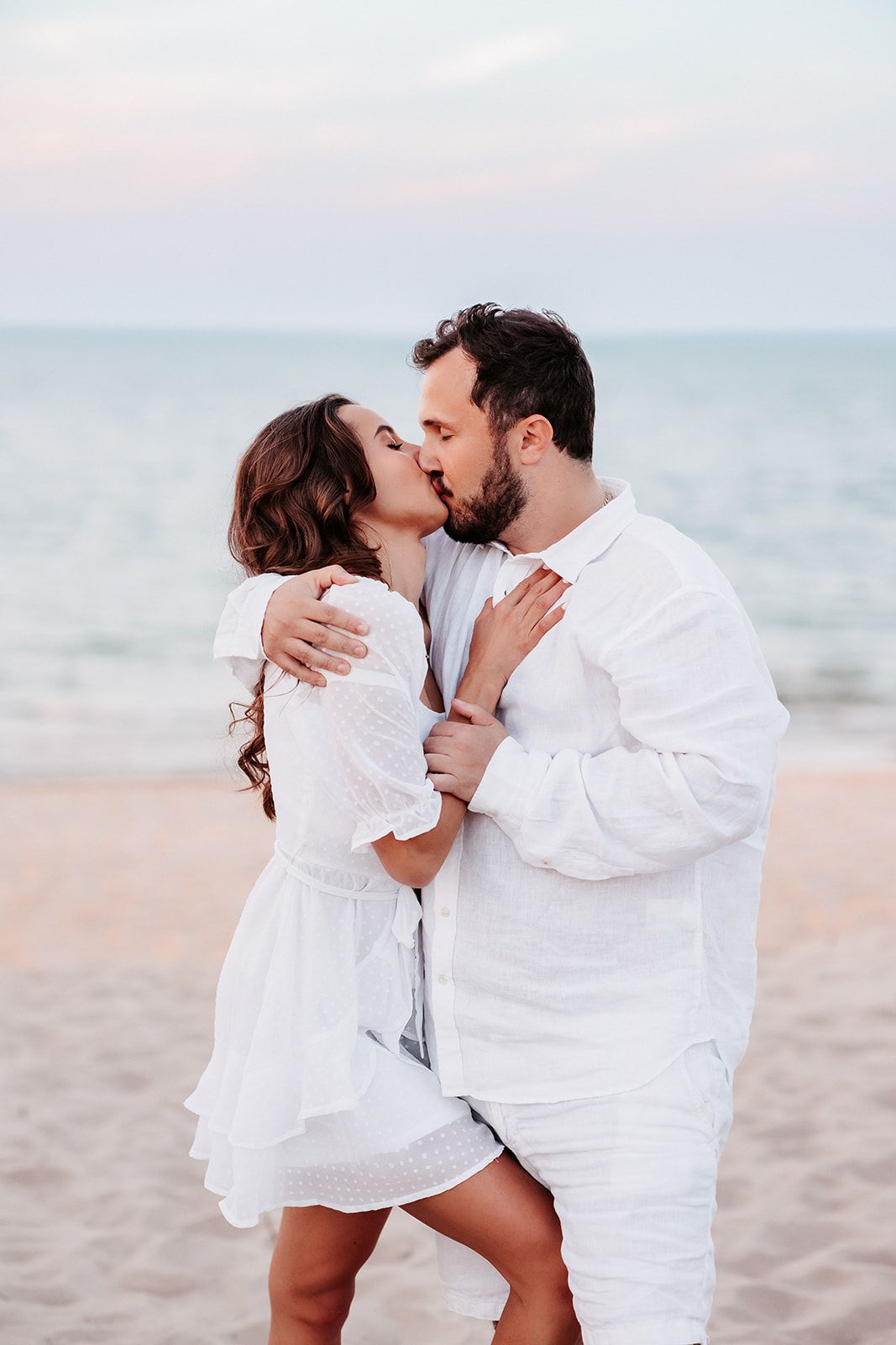 Couple in matching white outfits sharing a passionate kiss on the beach near the ocean.