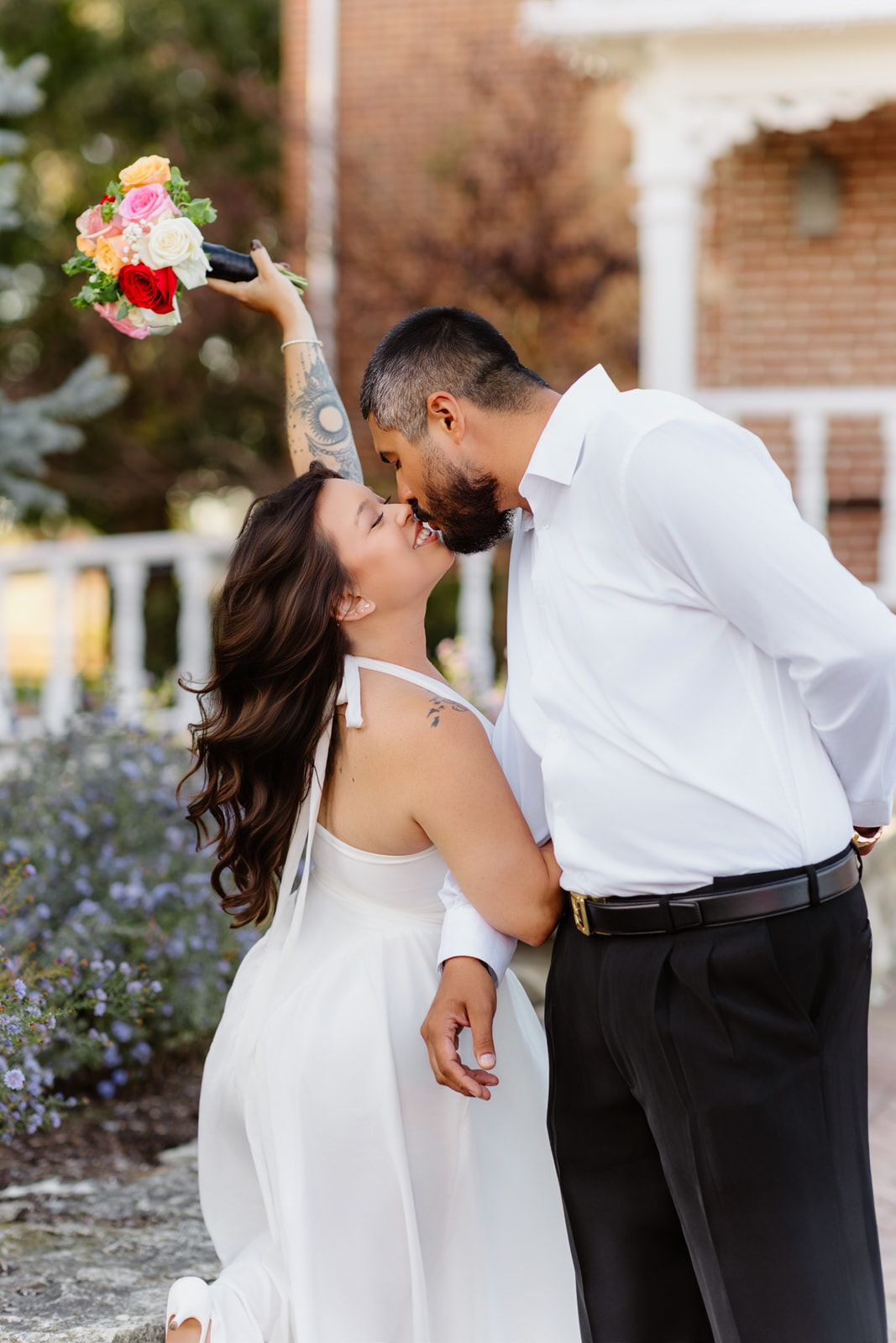 Bride and groom share a romantic kiss outdoors, bride holding colorful bouquet aloft in white halter dress.