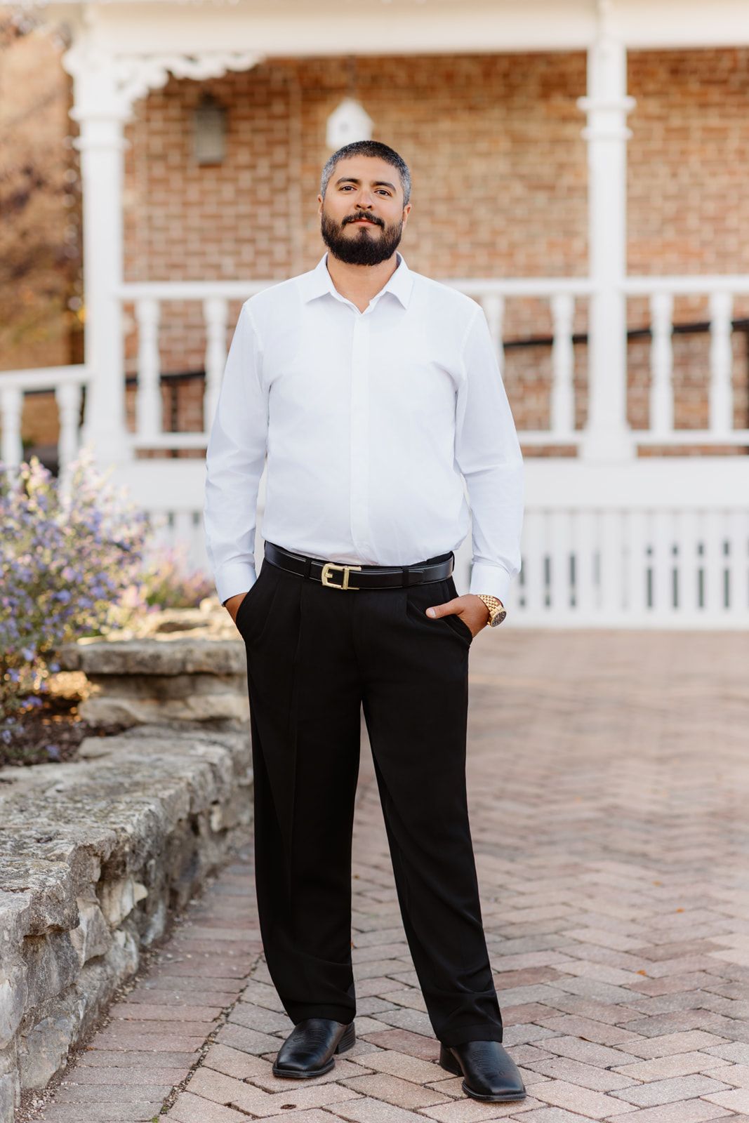 Groom in white dress shirt and black pants stands confidently outside a brick building with white railings.