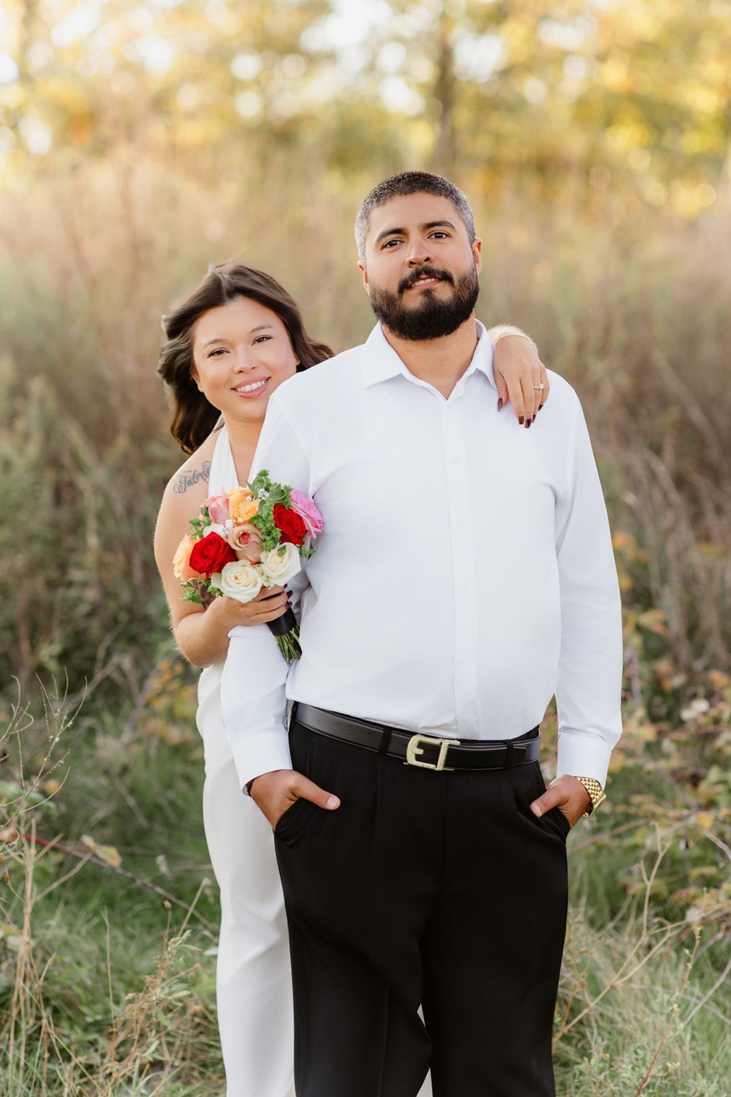 Bride and groom pose outdoors in white wedding attire, she holds a colorful bouquet while embracing him from behind.