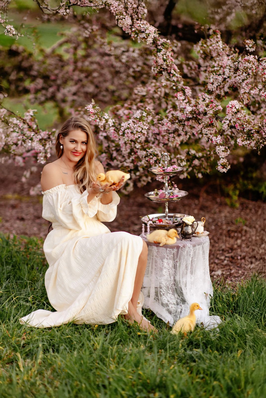 Smiling blonde woman in white dress holding pastries near tiered stand under pink blossoming tree.