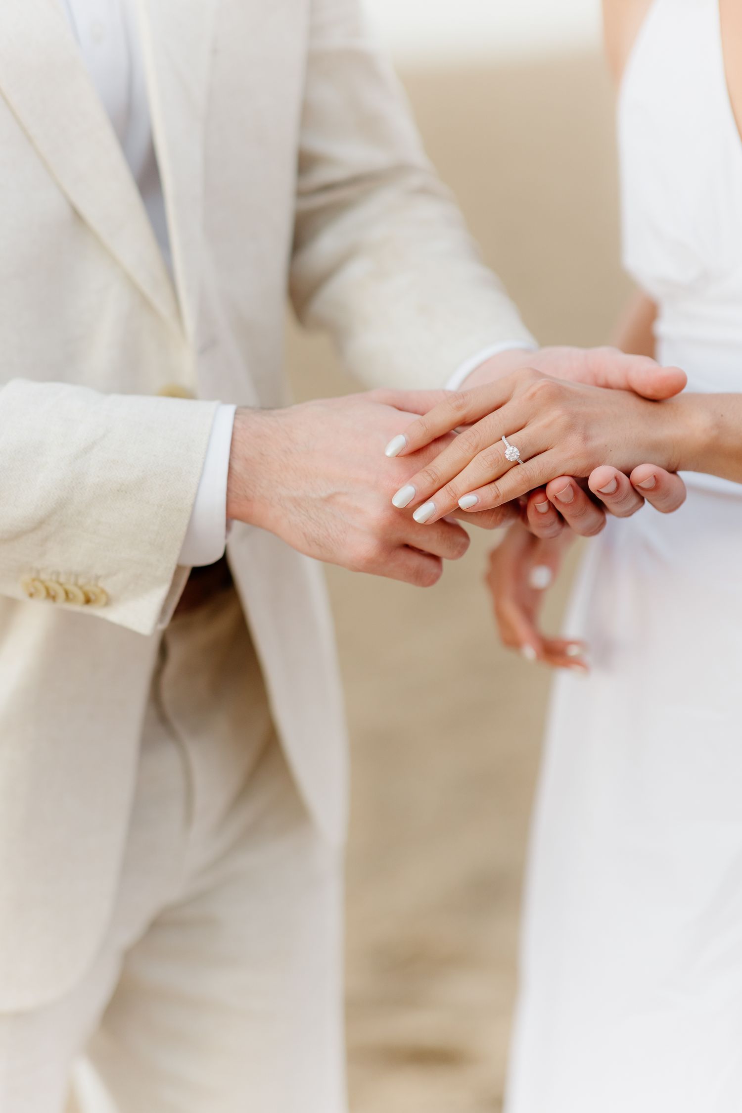 Close-up of bride and groom holding hands, showcasing elegant diamond engagement ring, wearing white and beige wedding attire.
