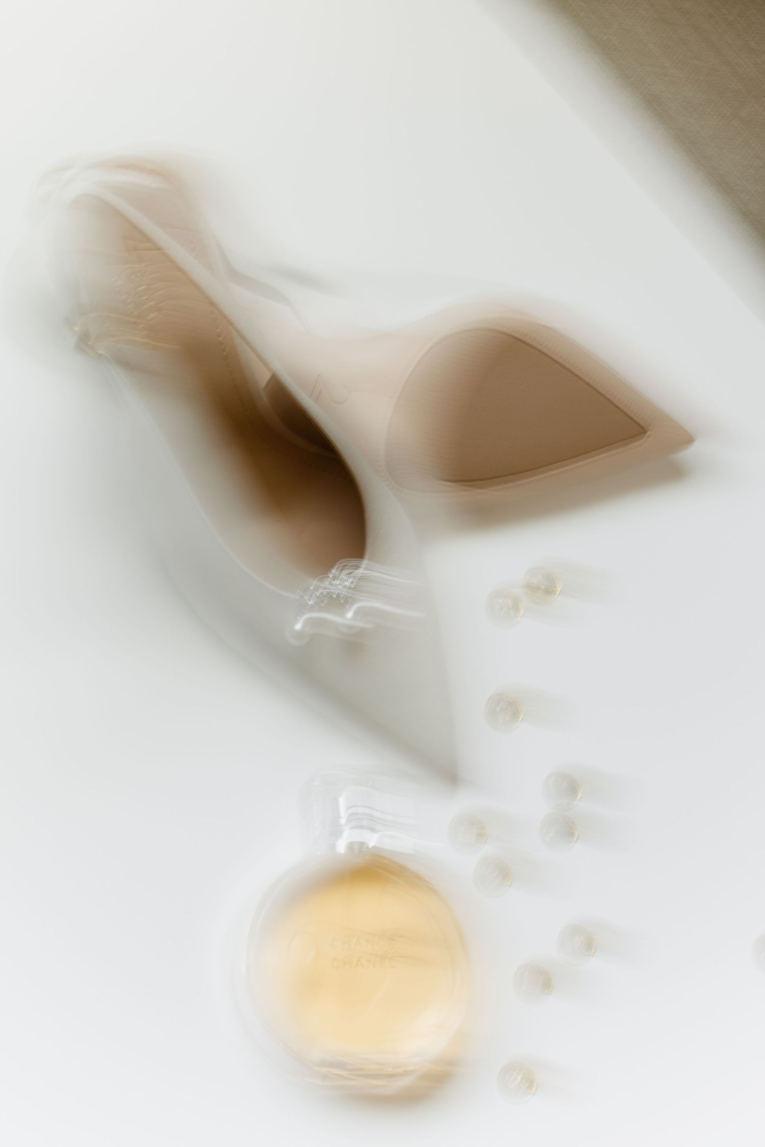 Artistic blurred motion shot of nude high heel shoes with pearls and perfume on white surface.