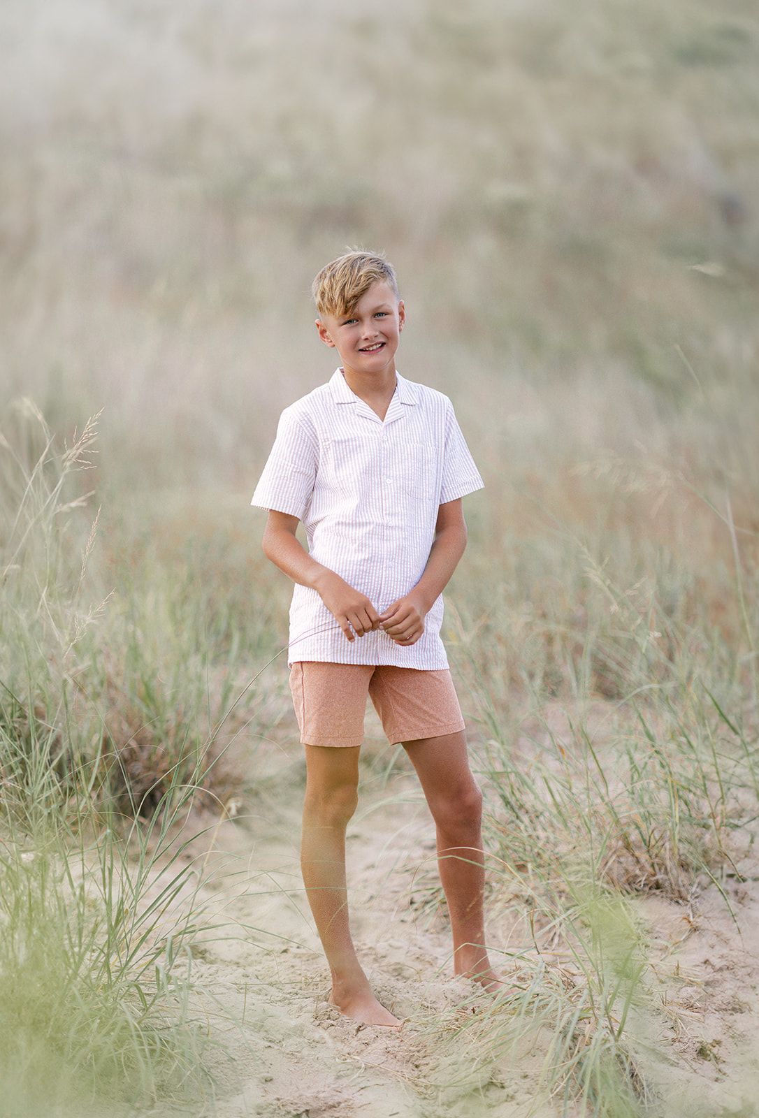Boy in white shirt and peach shorts standing barefoot on sandy beach dunes with tall grass background.