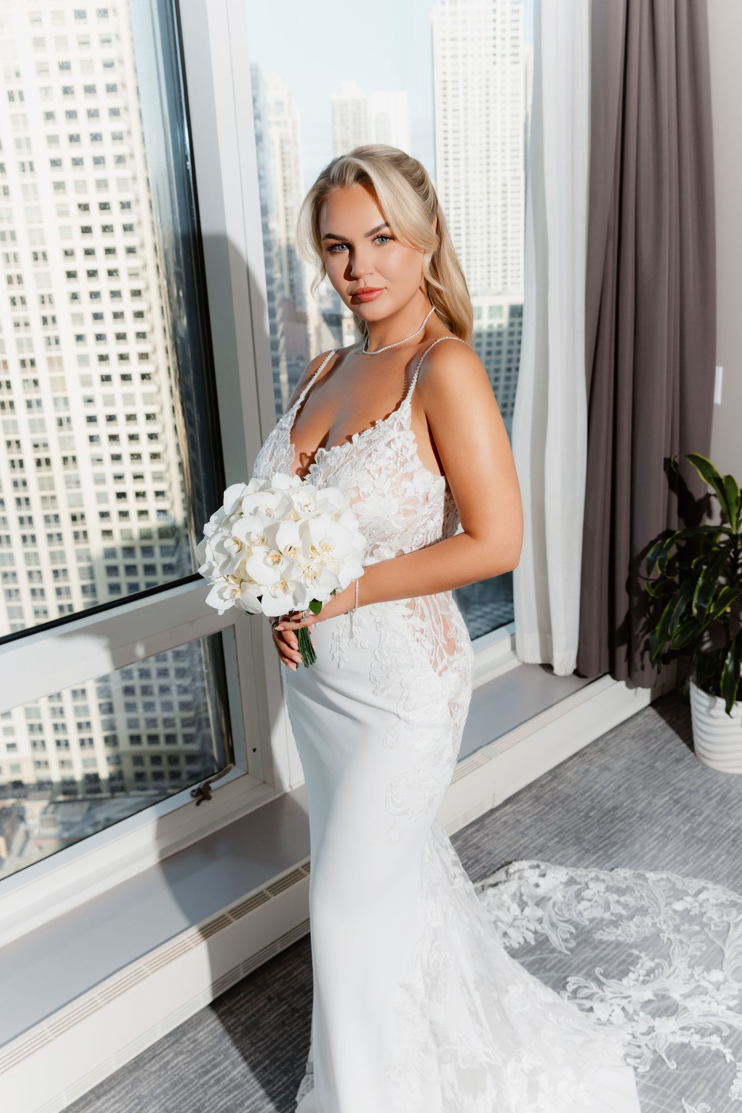 Bride in lace mermaid wedding gown holding white orchid bouquet by floor-to-ceiling city view windows.