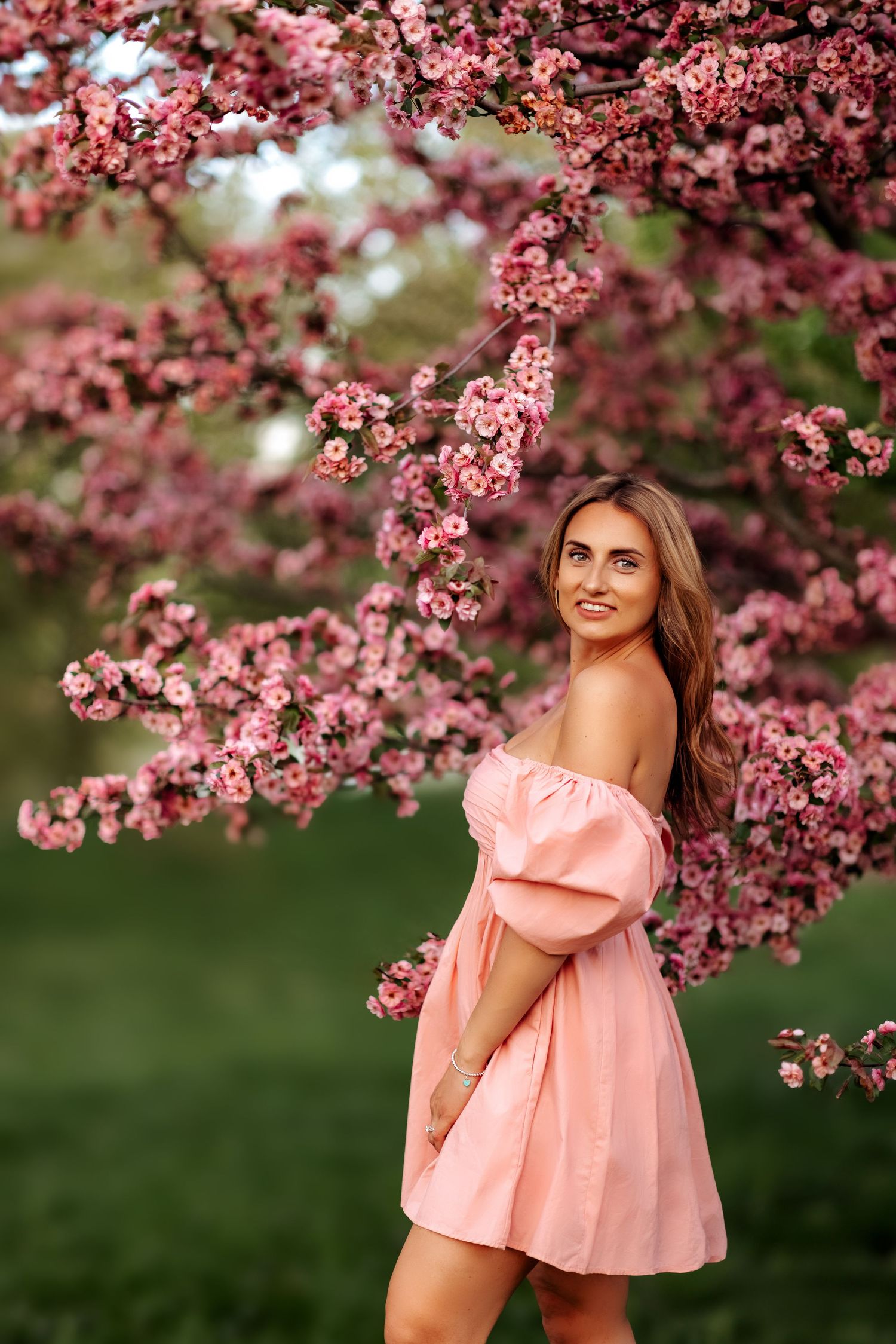 Beautiful woman in pink off-shoulder mini dress posing under vibrant pink cherry blossom tree.