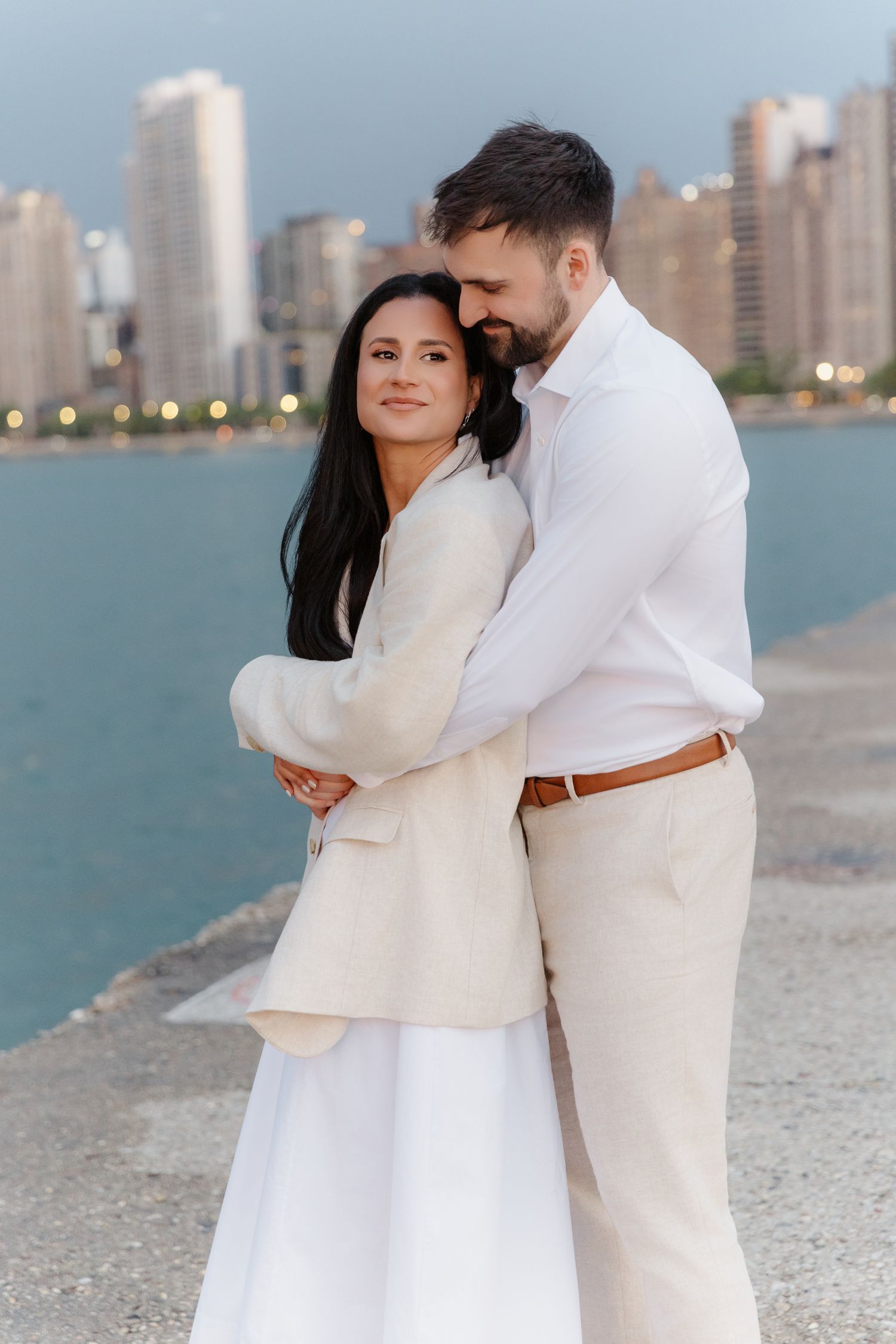 Couple cuddling on Chicago waterfront pier at dusk with glowing city skyline behind them.