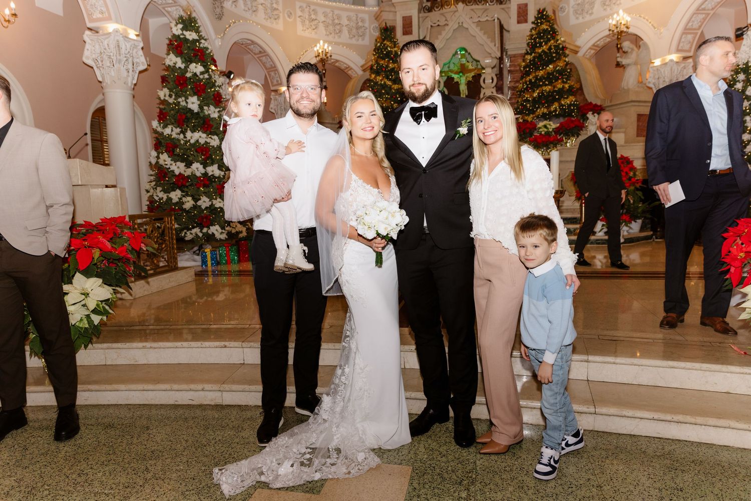 Bride in lace gown poses with groom and family members including children in decorated church lobby after wedding.