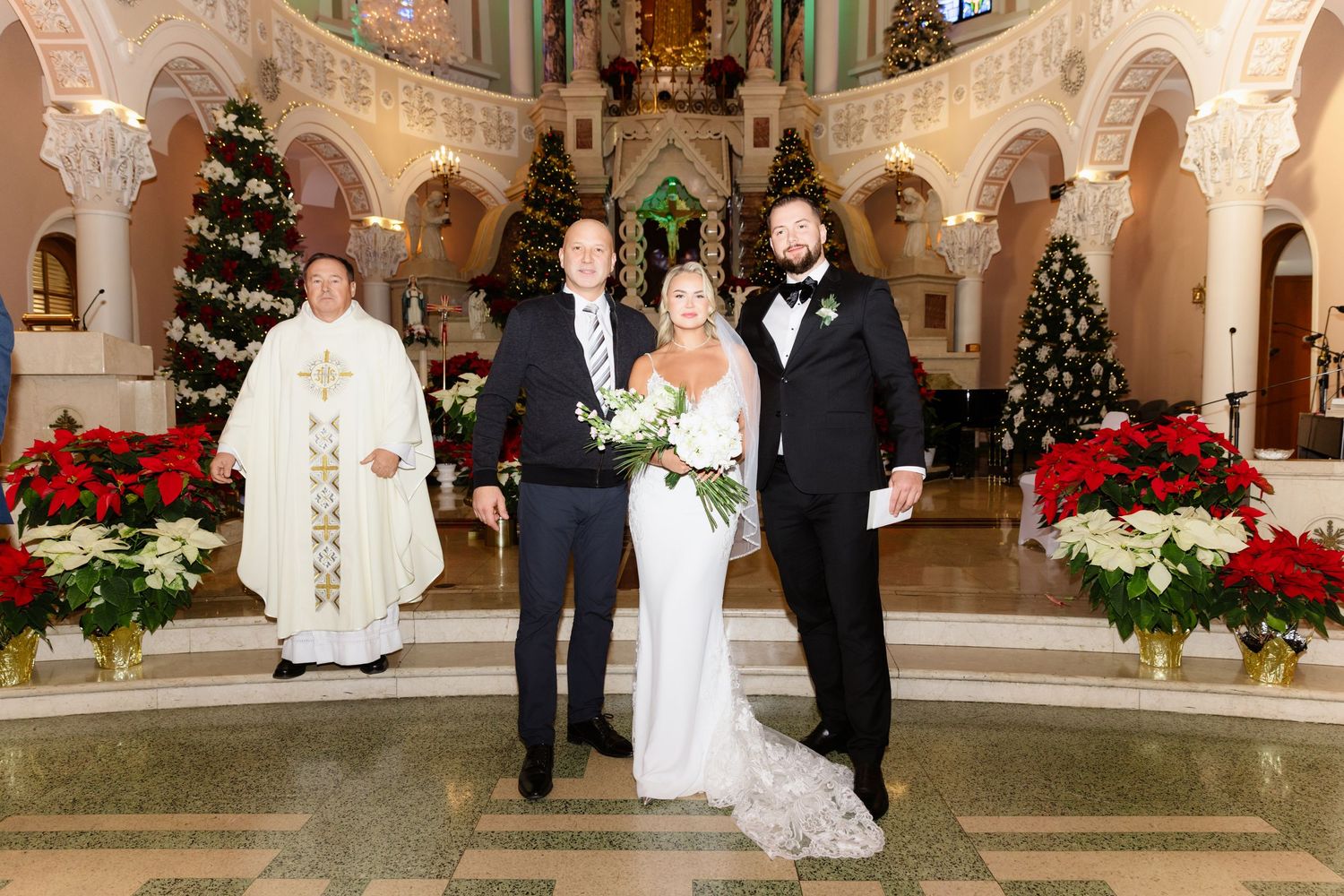 Newlyweds and priest posing at the altar of an elegant church adorned with Christmas trees and red poinsettia flowers.