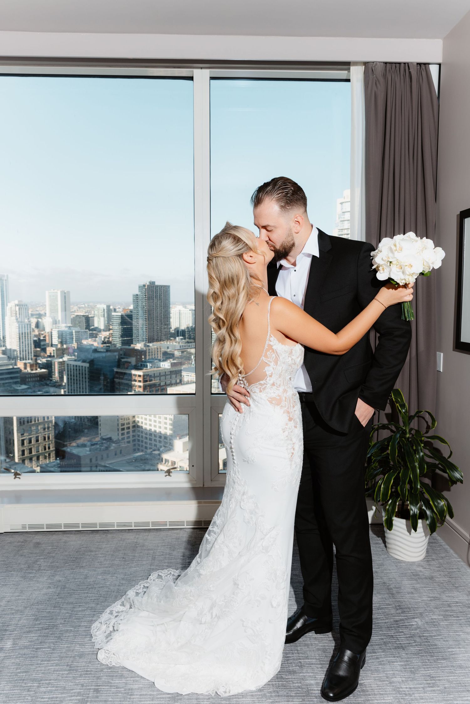 Couple shares a tender kiss in elegant hotel suite with stunning city skyline view behind them.