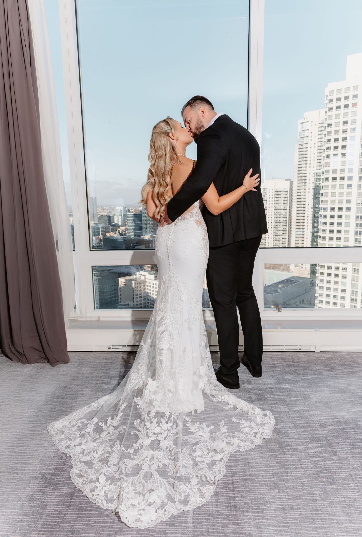 Bride in lace mermaid gown with cathedral train kissing groom in black tuxedo by city view window.